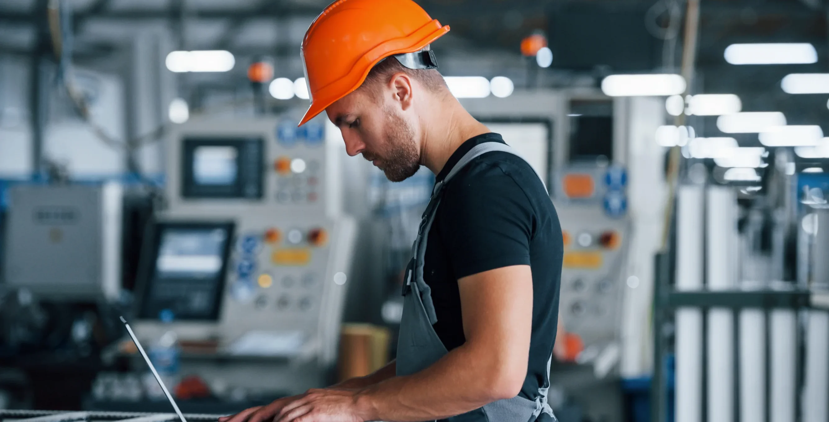 Industrial worker wearing a safety helmet using a laptop inside a manufacturing facility for manufacturing web development and digital operations.