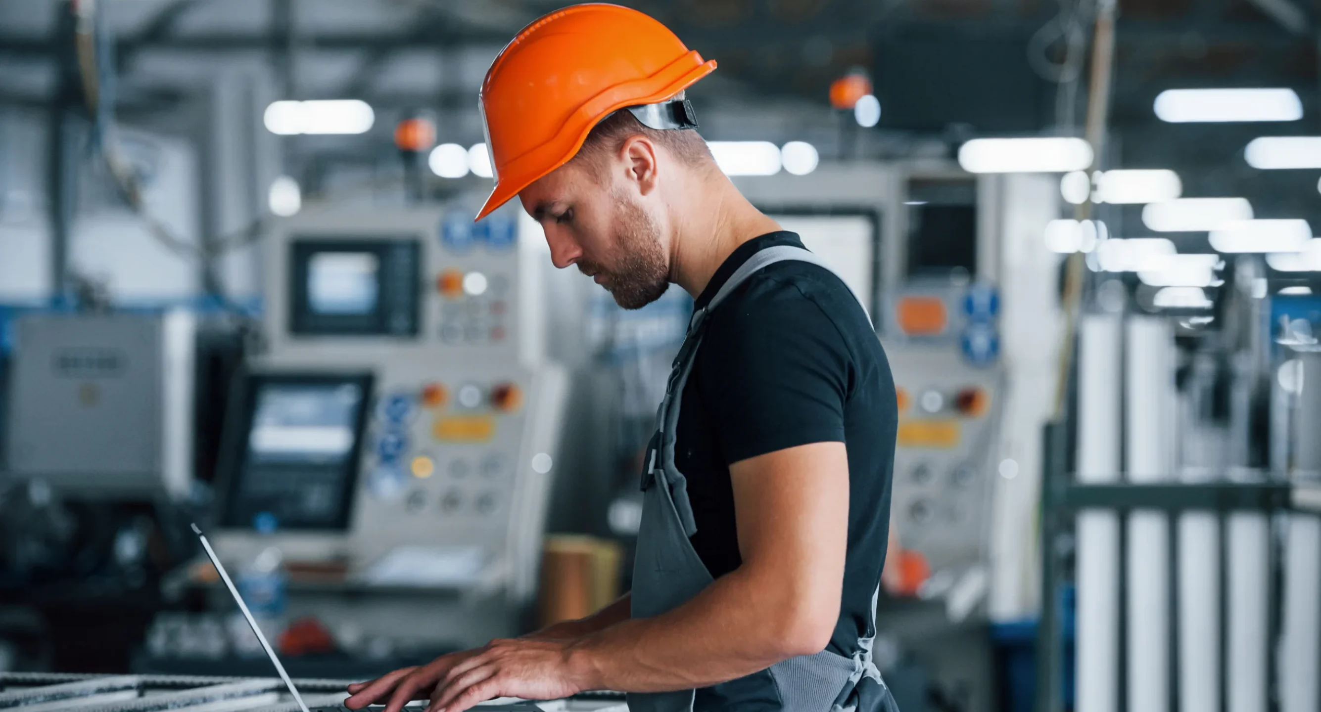 Industrial worker wearing a safety helmet using a laptop inside a manufacturing facility for manufacturing web development and digital operations.